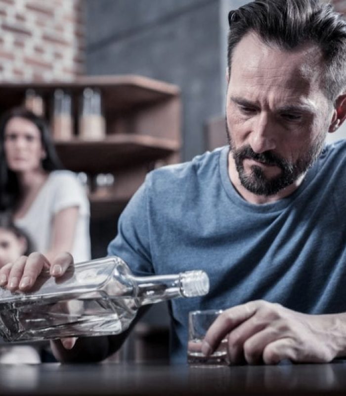 drug seeking behavior represented by man addicted to alcohol pours drink while wife watches sadly in back